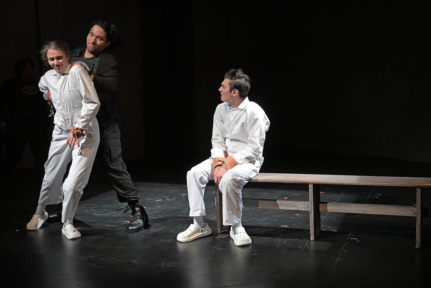 One student actor in a white jumpsuit watches as an actor playing a prison guard throws another actor in a white jumpsuit onto the stage.