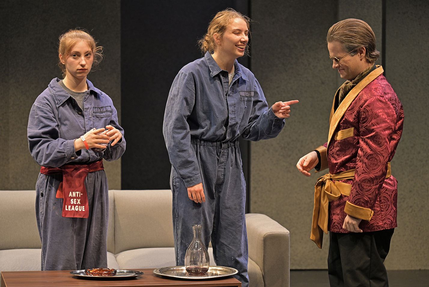 Two actors in blue laborer jumpsuits speak to a third actor in a lounge jacket during a scene in a play.