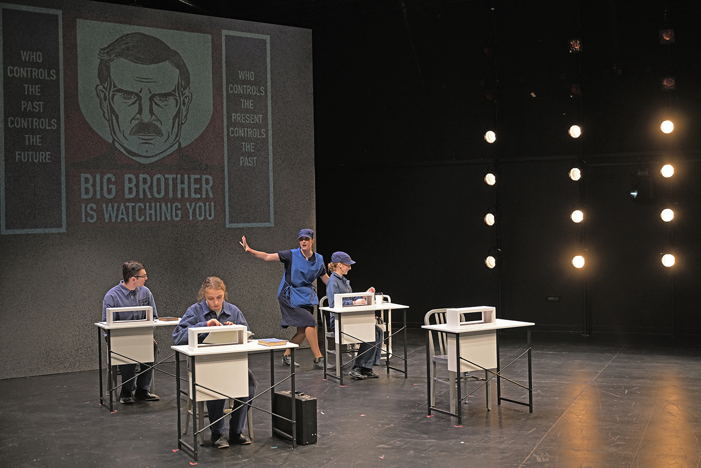 One actor in a blue jumpsuit walks through a room with three actors in blue jumpsuits sitting at white desks with an image of Big Brother projected on the wall behind them.