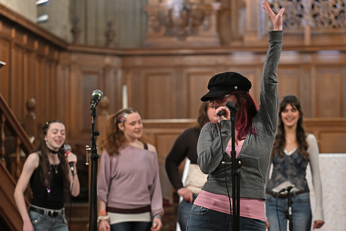 A soloist with a college a cappella group holds one hand up as she sings into a microphone with fellow singers behind.