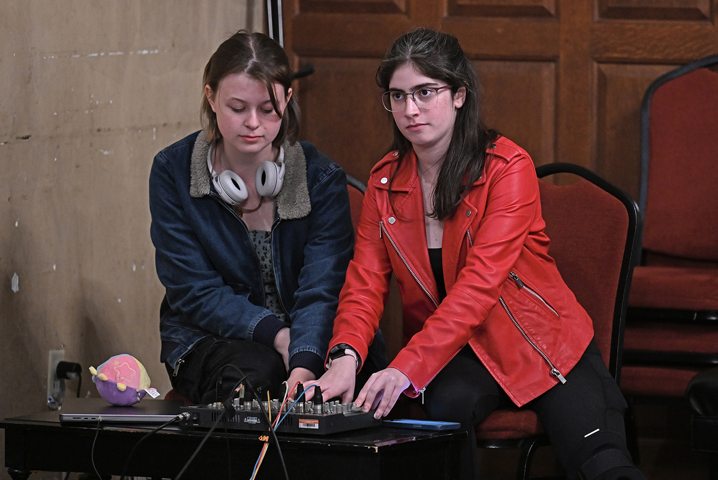 Two female college students, one in a red leather jacked, the other with headphones around her neck, work at a sound board during an a cappella concert.