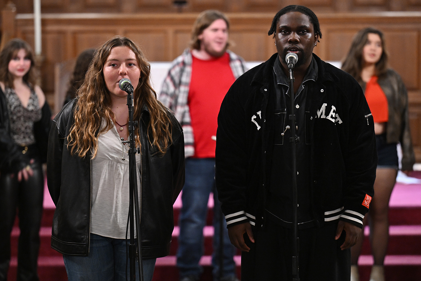 A pair of college a cappella singers, one female and one make, sing at two microphones out in front of their group.