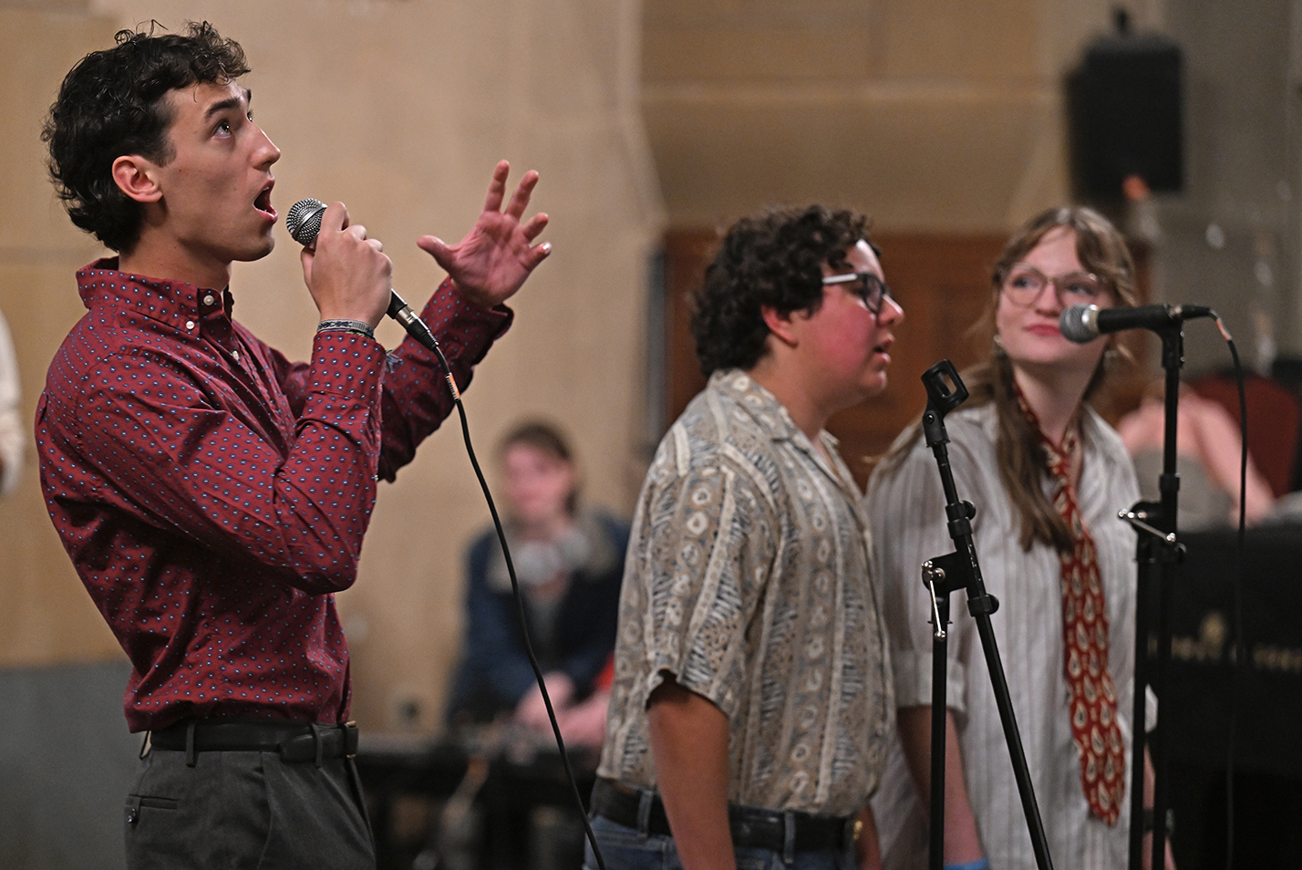 A male soloist with curly hair raises his left hand and looks toward the ceiling as he performs a solo with a college a cappella group.