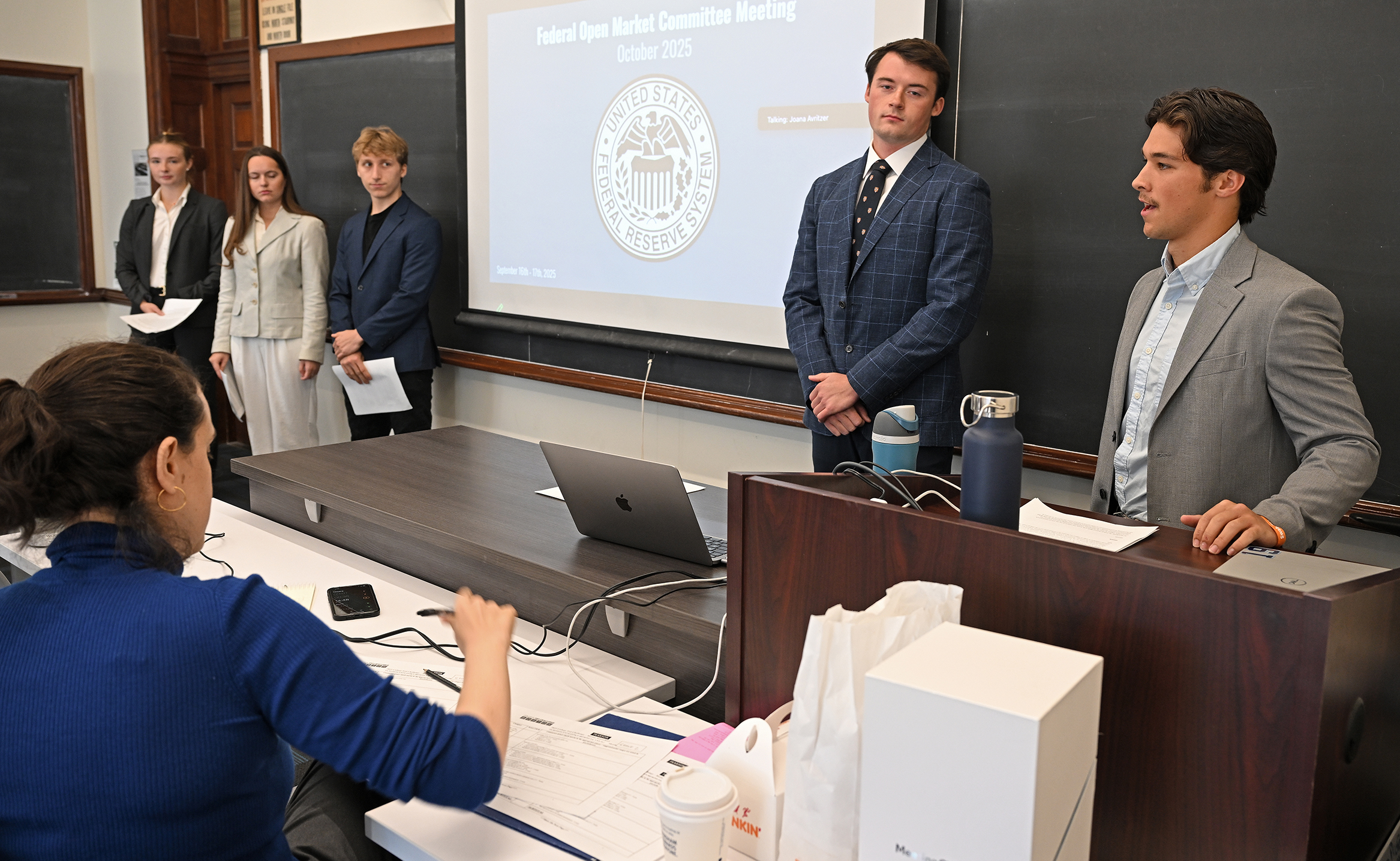 Five college students in business attire stand at the front of a classroom and make a presentation.