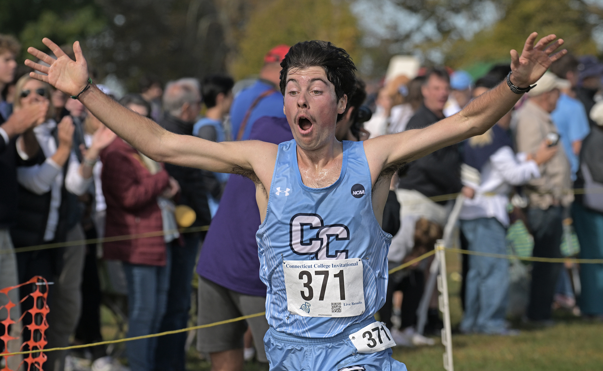 A collegiate cross-country runner in light blue uniform and CC on the front with the bib number throws his hands up in the air with a surprised look on his face as he crosses the finish line of a race.