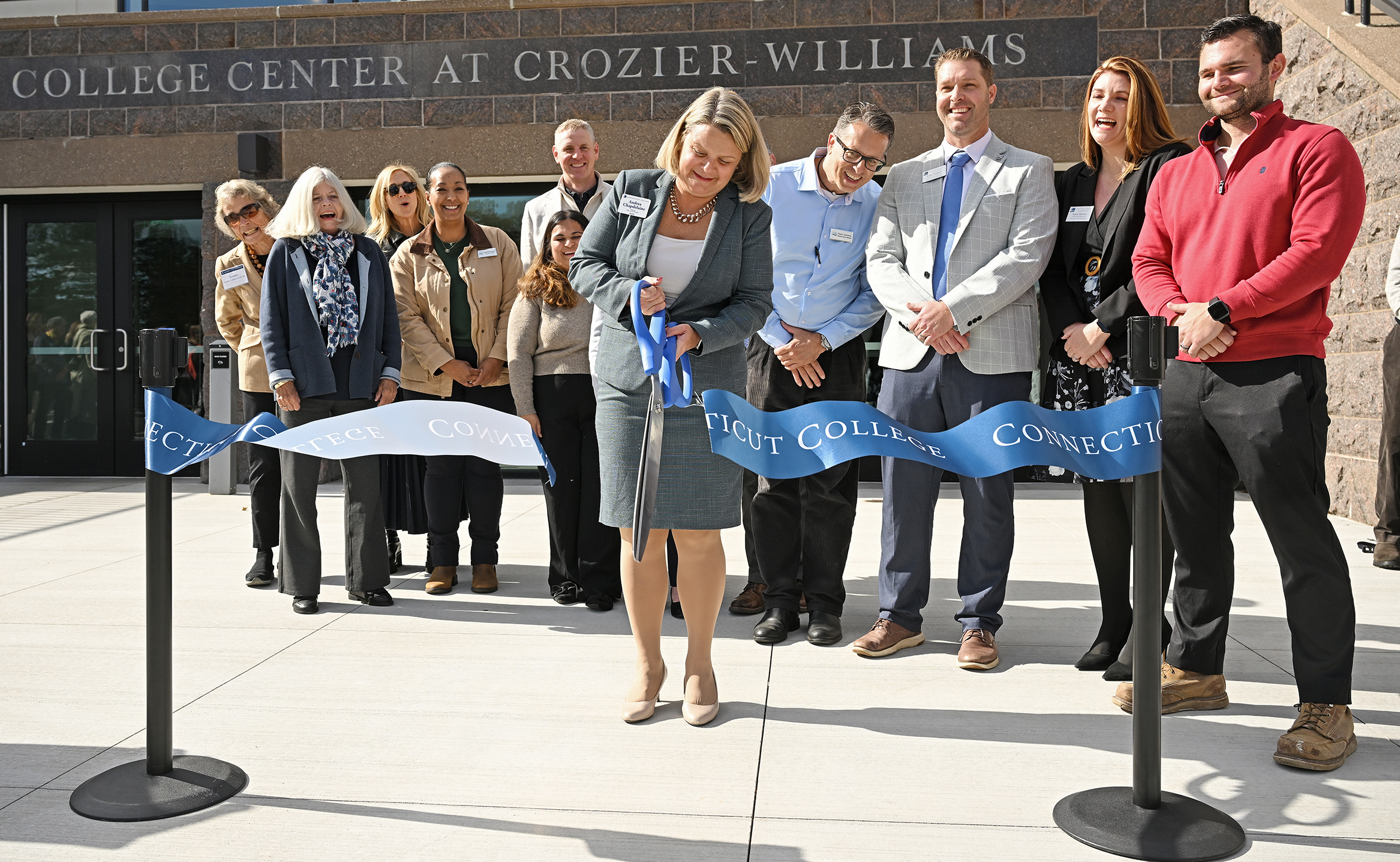 A college president cuts a giant ribbon with giant scissors surrounded by various VIP's during a building dedication.