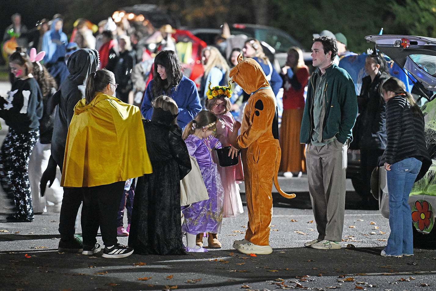 Children and college students in halloween costumes interact during a trunk or treat event in a parking lot after dark.