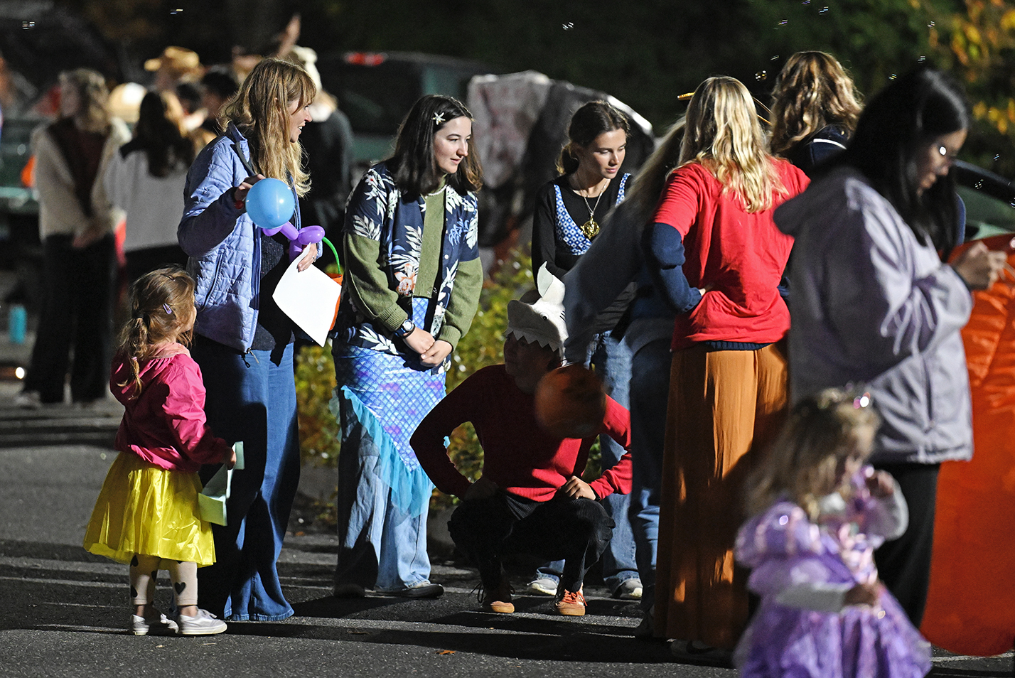 Children and college students in halloween costumes interact during a trunk or treat event in a parking lot after dark.