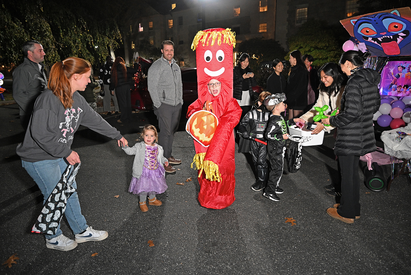 Children and college students in halloween costumes interact during a trunk or treat event in a parking lot after dark.