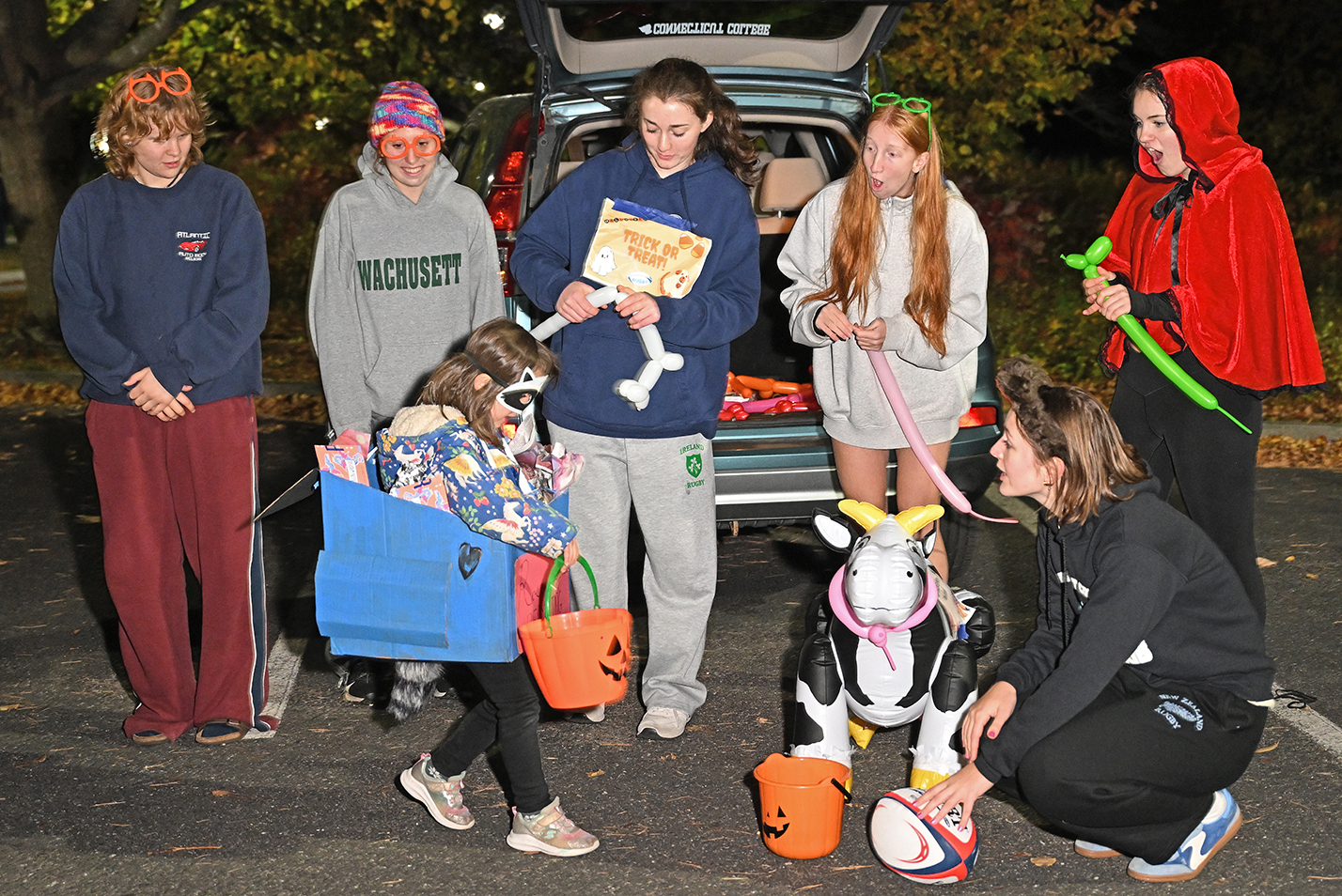 Children and college students in halloween costumes interact during a trunk or treat event in a parking lot after dark.
