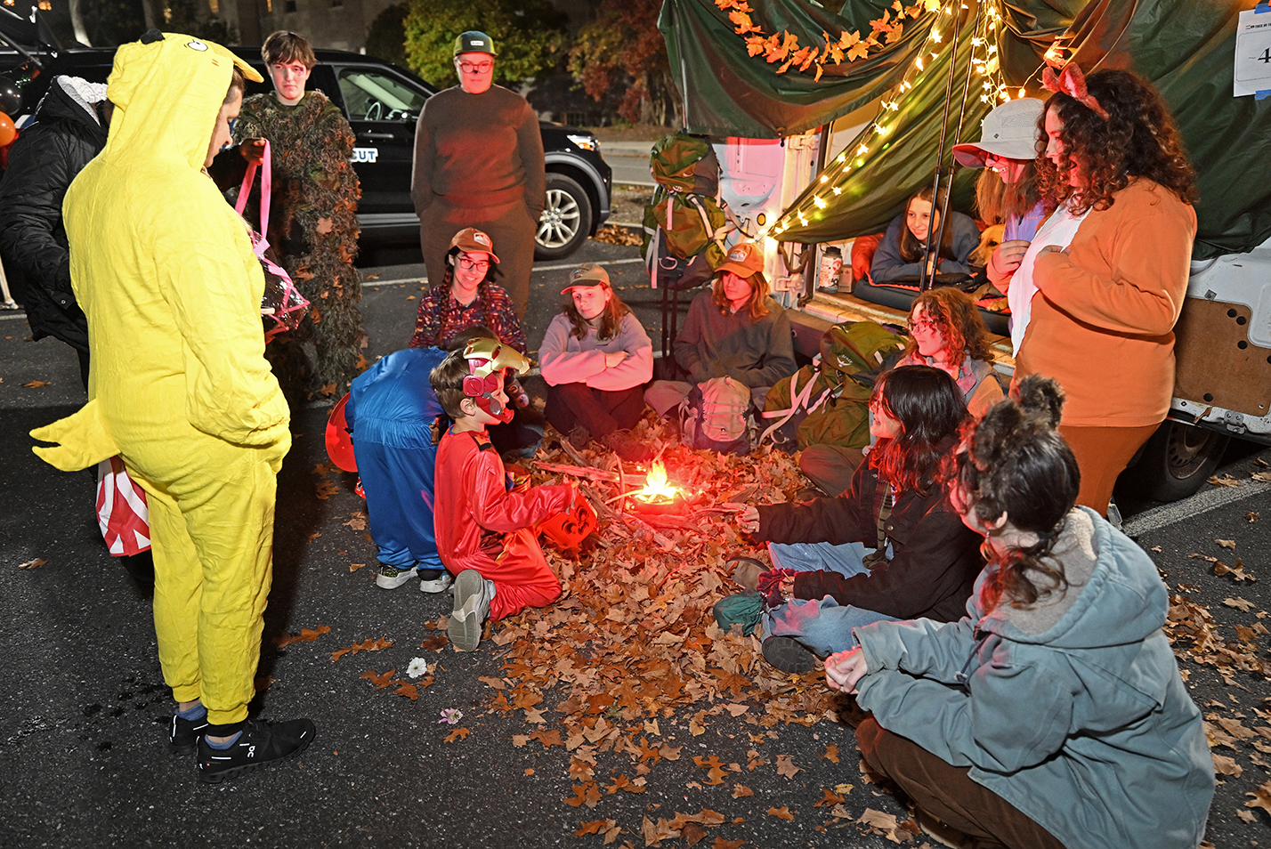 Children and college students in halloween costumes interact during a trunk or treat event in a parking lot after dark.