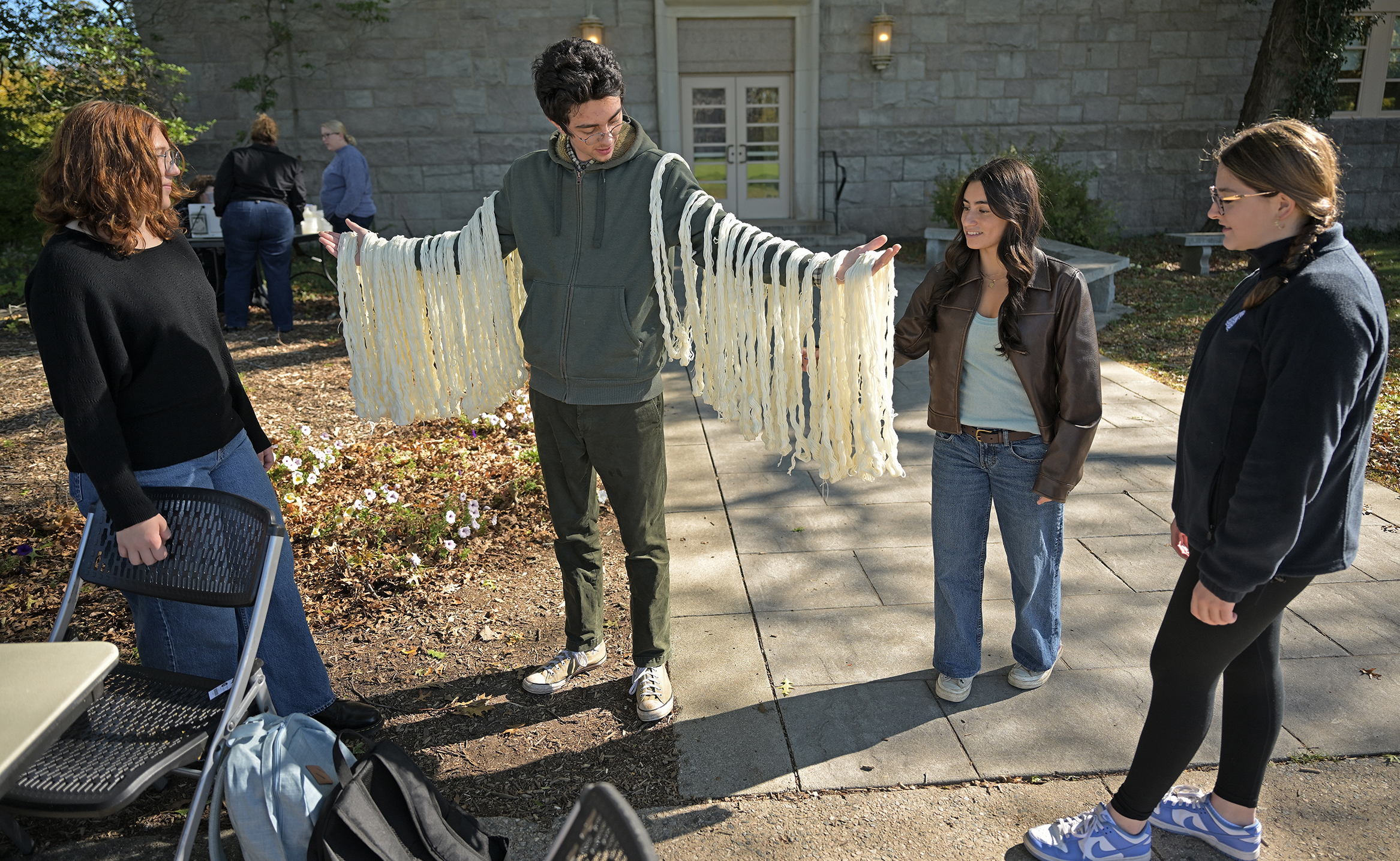 A male college student in a hoodie sweatshirt holds his arms out with loops of wool yarn draped over them as three female students look on.