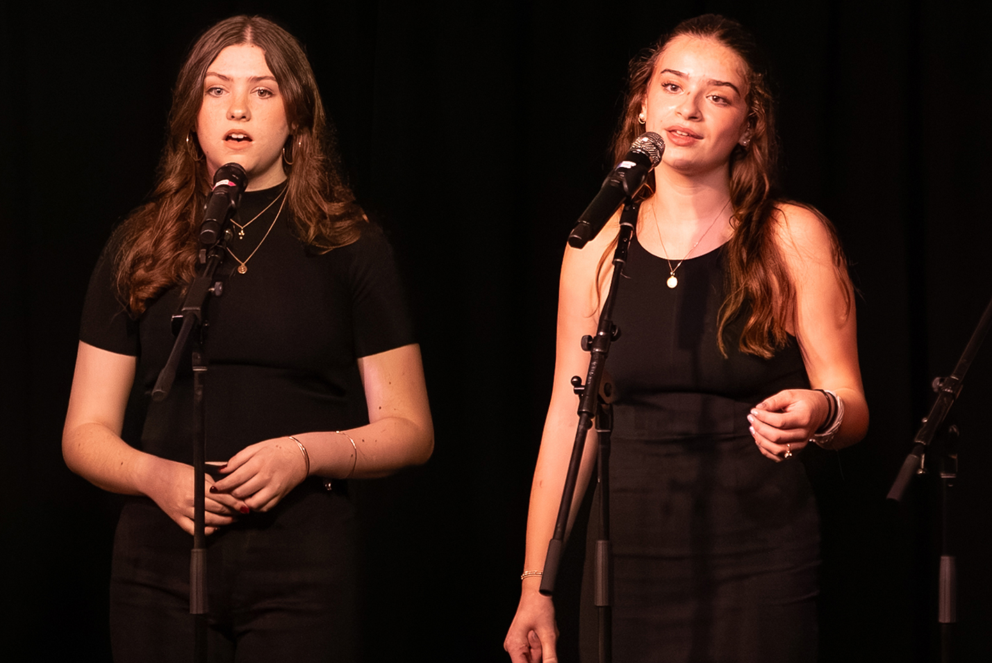 Two members of an all-female college a cappella group in black dresses performs on stage.