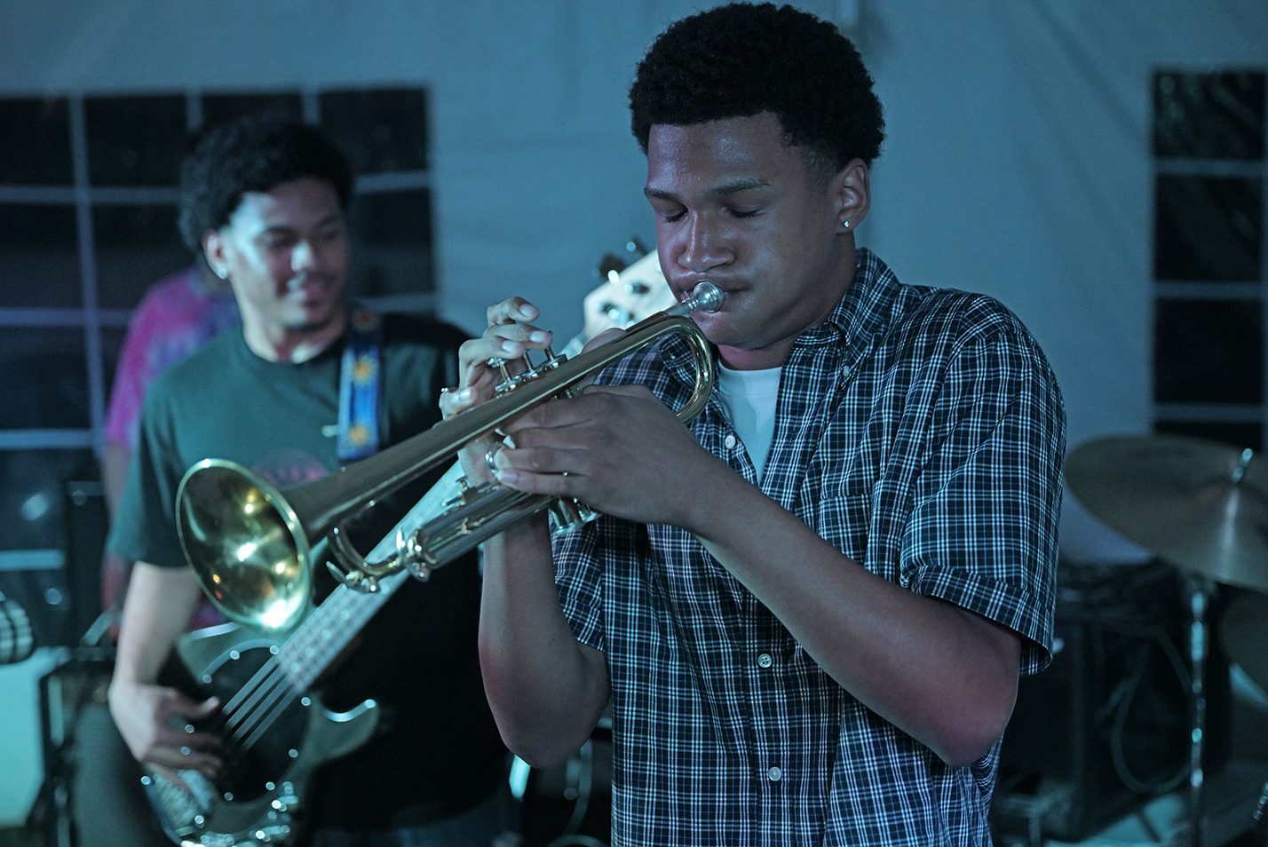 A student musician plays the trumpet during a rock band concert on a low stage under a tent.