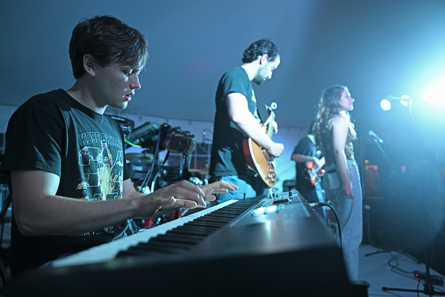 A student musician plays an electronic keyboard with a band on a stage under a tent.
