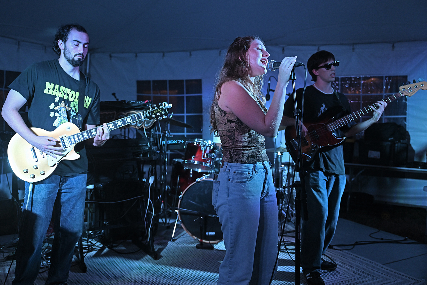A female student sings accompanied by male students on guitars on a stage under a tent.