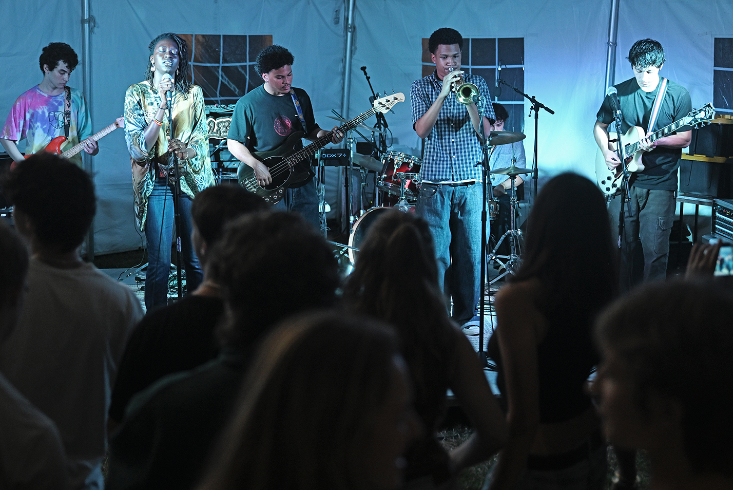 A six-piece student band performs on a low stage under a tent.