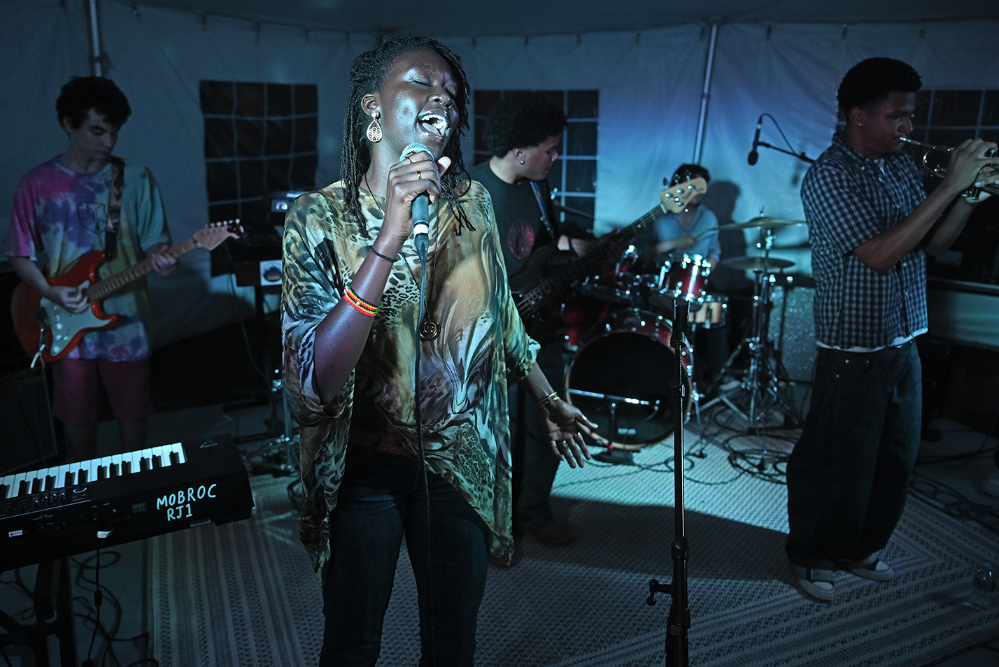 A student singer in a frilly patterned shawl performs with a band on a low stage under a tent.