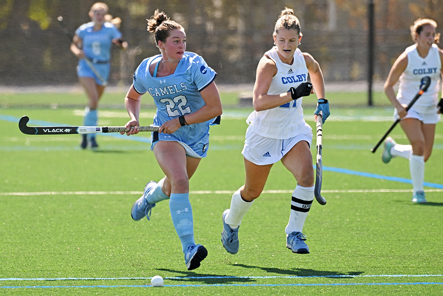 A field hockey player in light blue races up the field with the ball pursued by a player in white