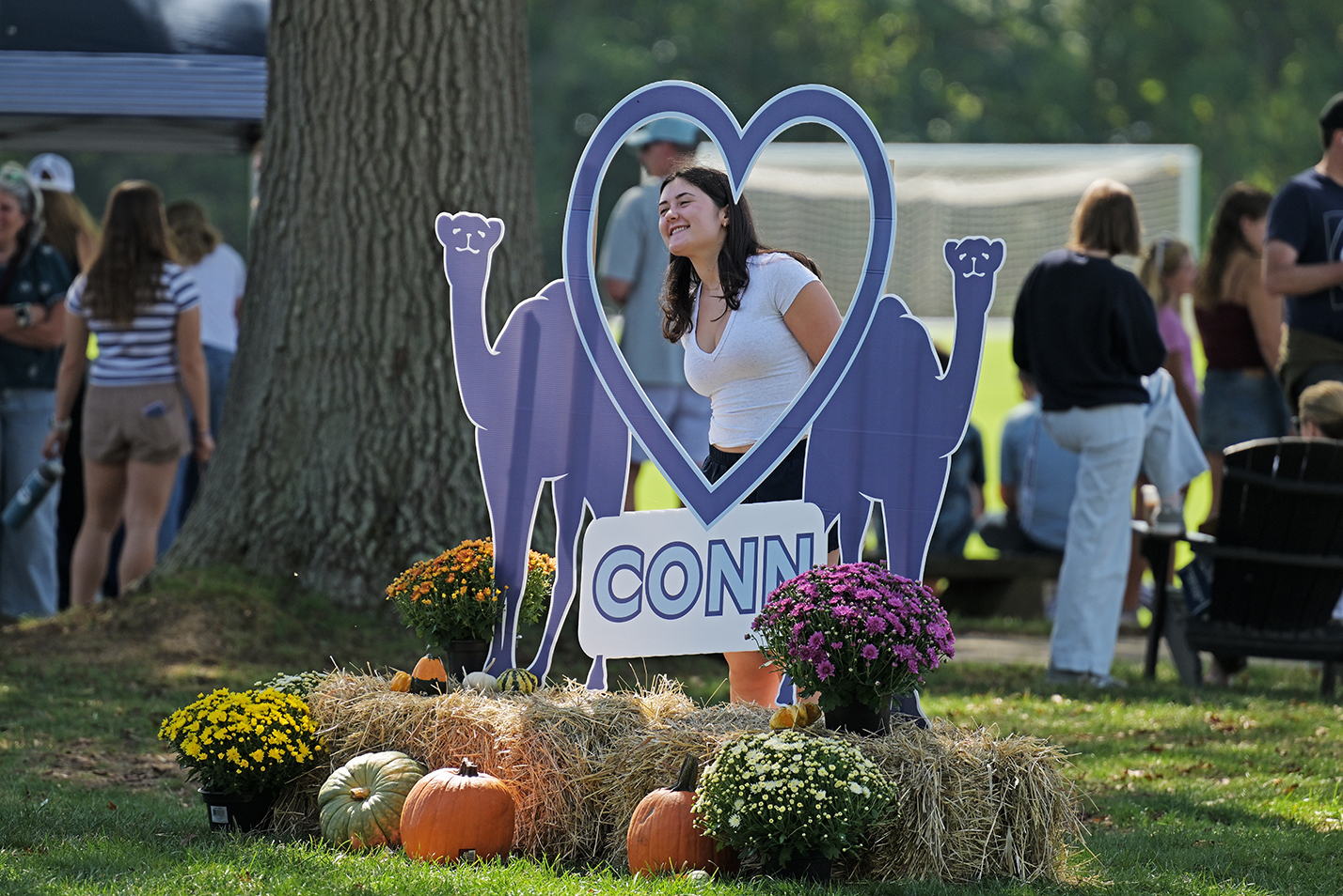 A college student poses in a large photo frame shaped like a heart flanked by two silhouettes of camels all atop a display of hay bales and pumpkins.