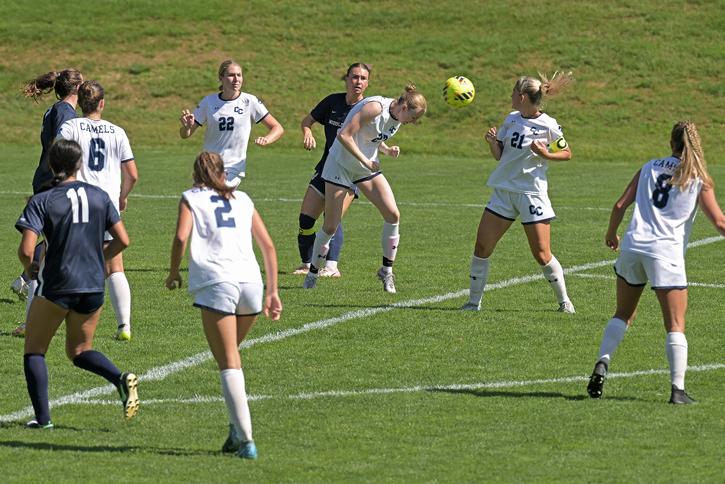 A female soccer player heads the ball surrounded by teammates and opponents on a natural grass pitch.