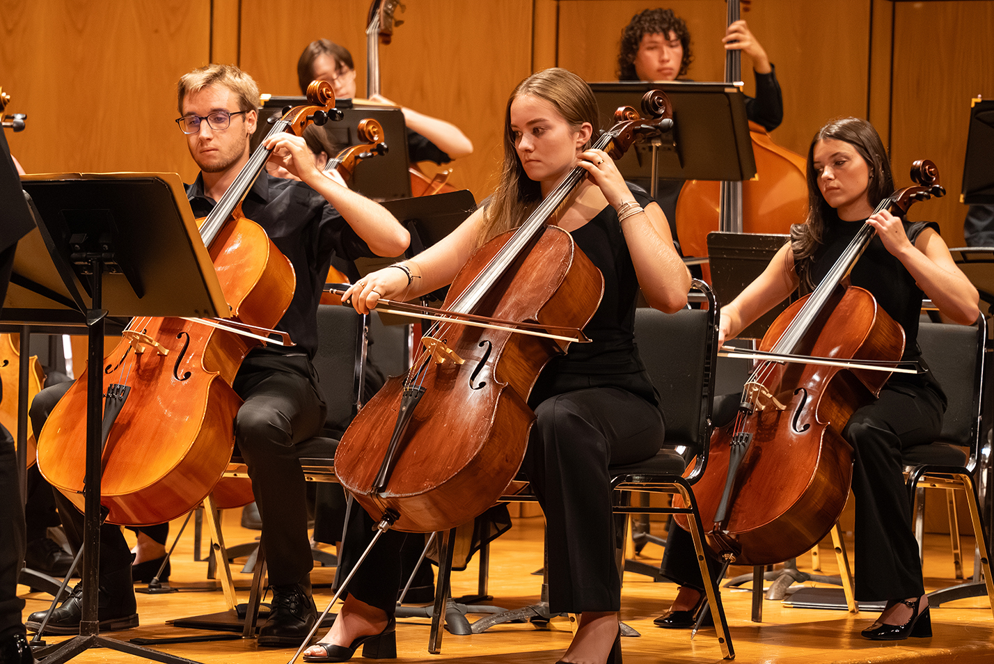 Student musicians on cello and bass perform with an orchestra on stage.