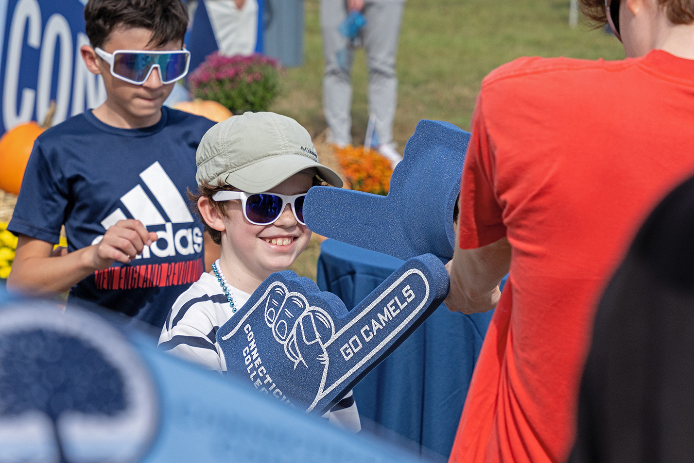 A child in hat and sunglasses waves a blue foam finger at another person.
