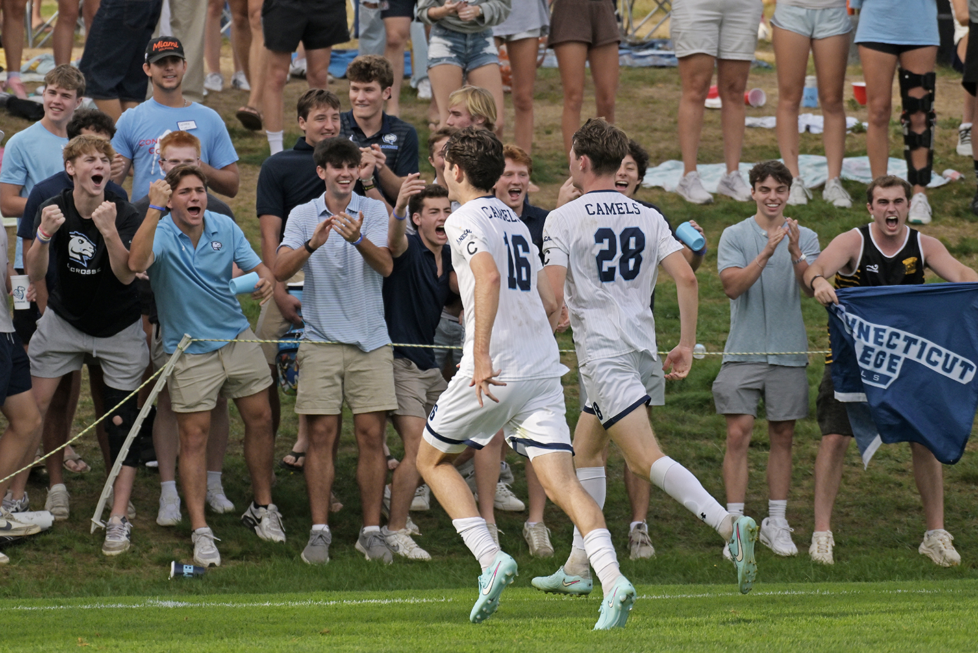 Two male collegiate soccer players in white uniforms run to the sideline of the pitch to join celebrating fans.