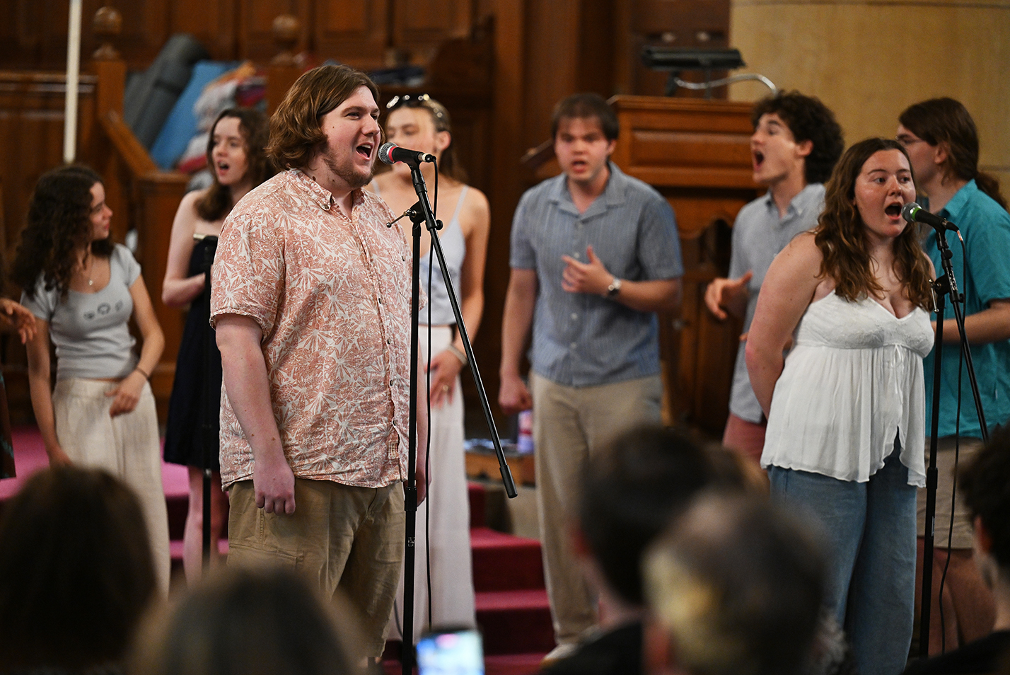 Two a cappella singers, one male, one female, stand and sing at microphones in front of a mixed-gender a cappella group.