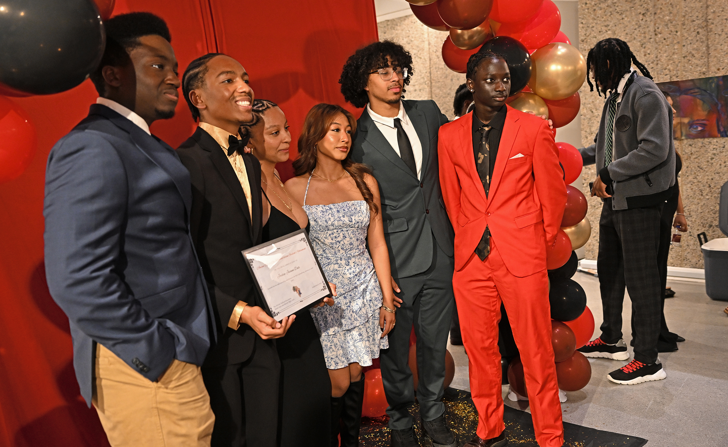 A group of students, male and female, of color dressed in suits and dresses, with one holding an award certificate, pose for a photo under an arch or red and gold balloons.