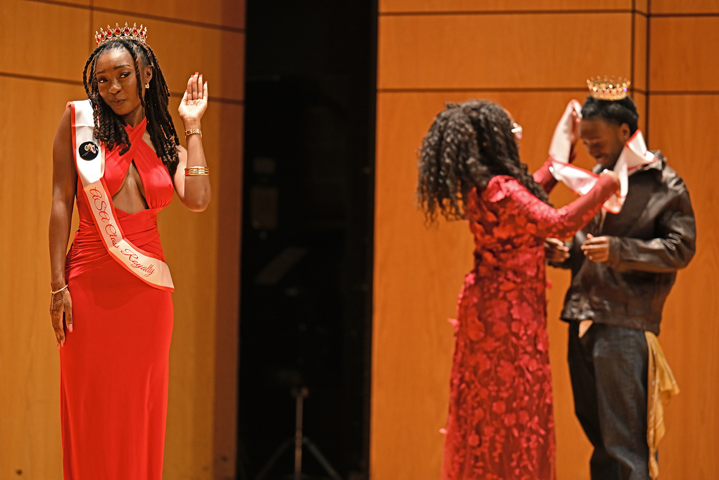 A young black woman in a long red dress with a crown and sash gives a princess wave while another women in a red dress places a sash around the shoulders of a young black man wearing a crown.
