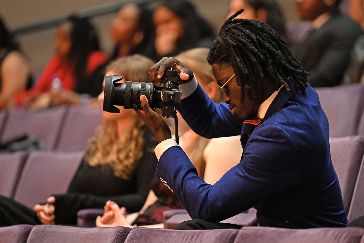 A young black man in a blue sportcoat and red bowtie takes photos with a large camera while seated in an auditorium.