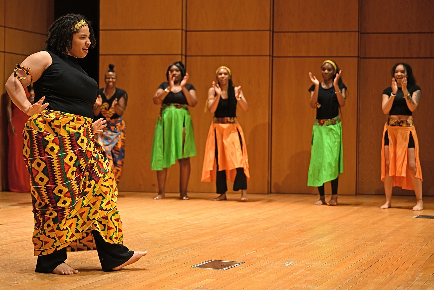 A young woman in colorful west African costume dances on a stage with other similarly attired young women dancing in the background.
