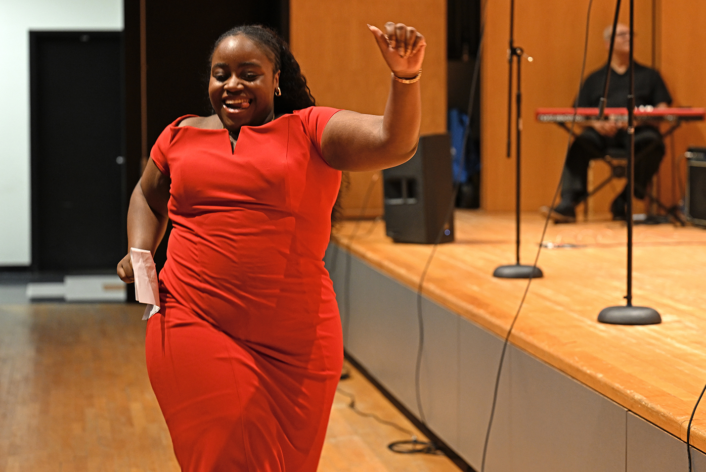 A young black woman in a red dress struts across the auditorium in front of a stage.