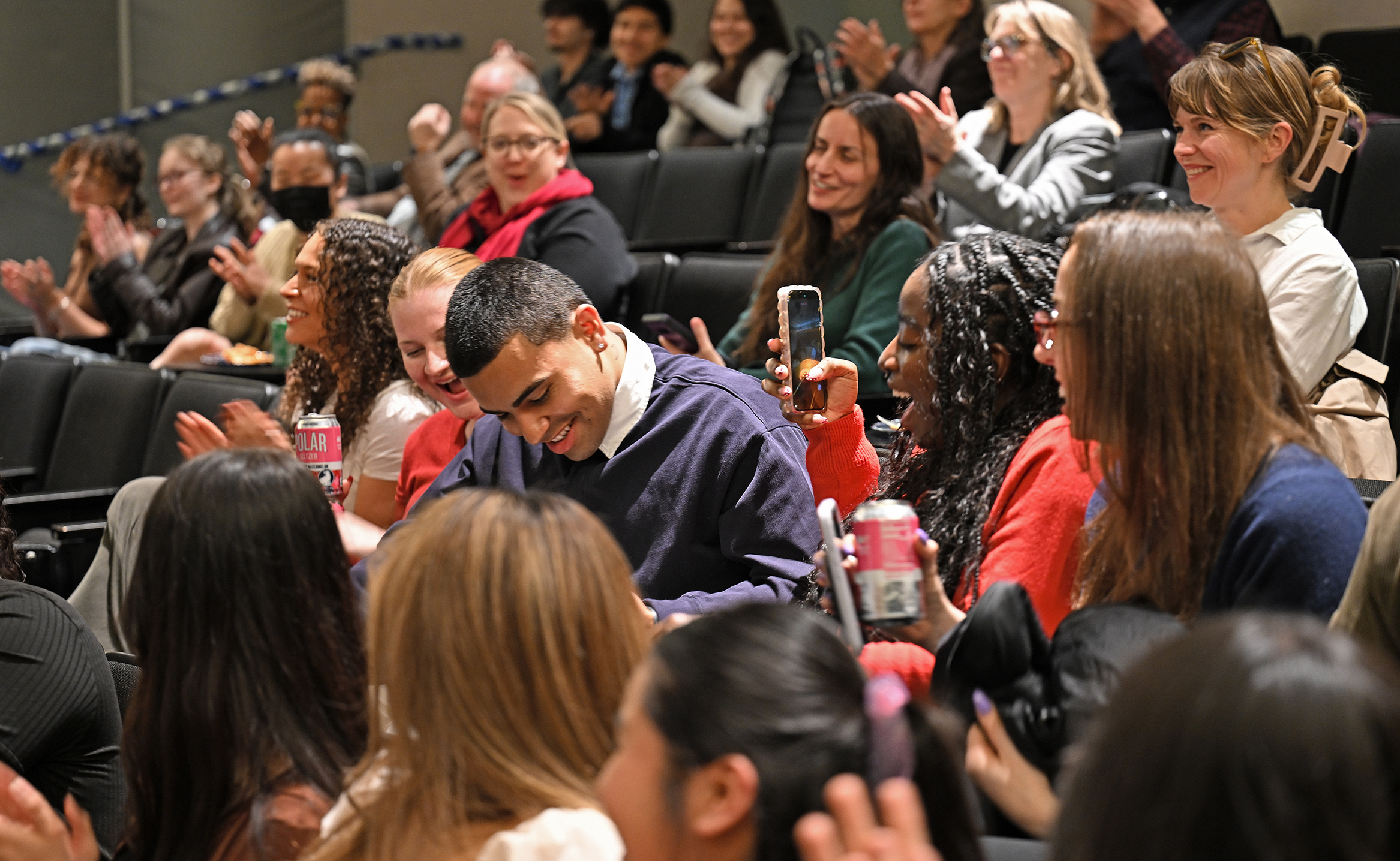 A male student bows his head surrounded by fellow students applauding and taking photos seated in an auditorium.