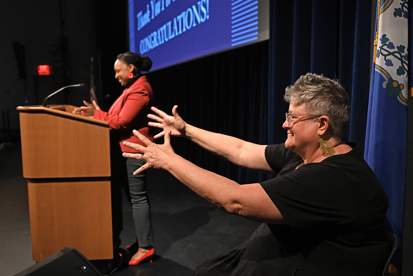 A sign language interpreter gestures while providing translation for a speaker standing at a podium.