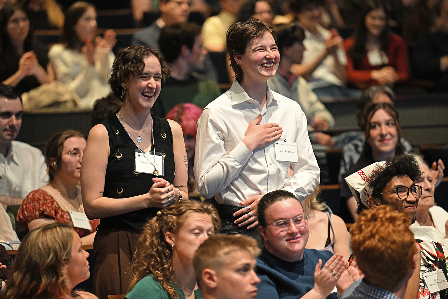 Two college students stand and smile broadly while surrounded by applauding classmates in an auditorium.