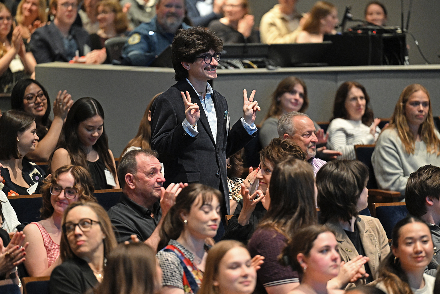 A male student with a mustache in a blue blazer gives two finger v-signs with each hand as he stands in an auditorium surrounded by applauding classmates.