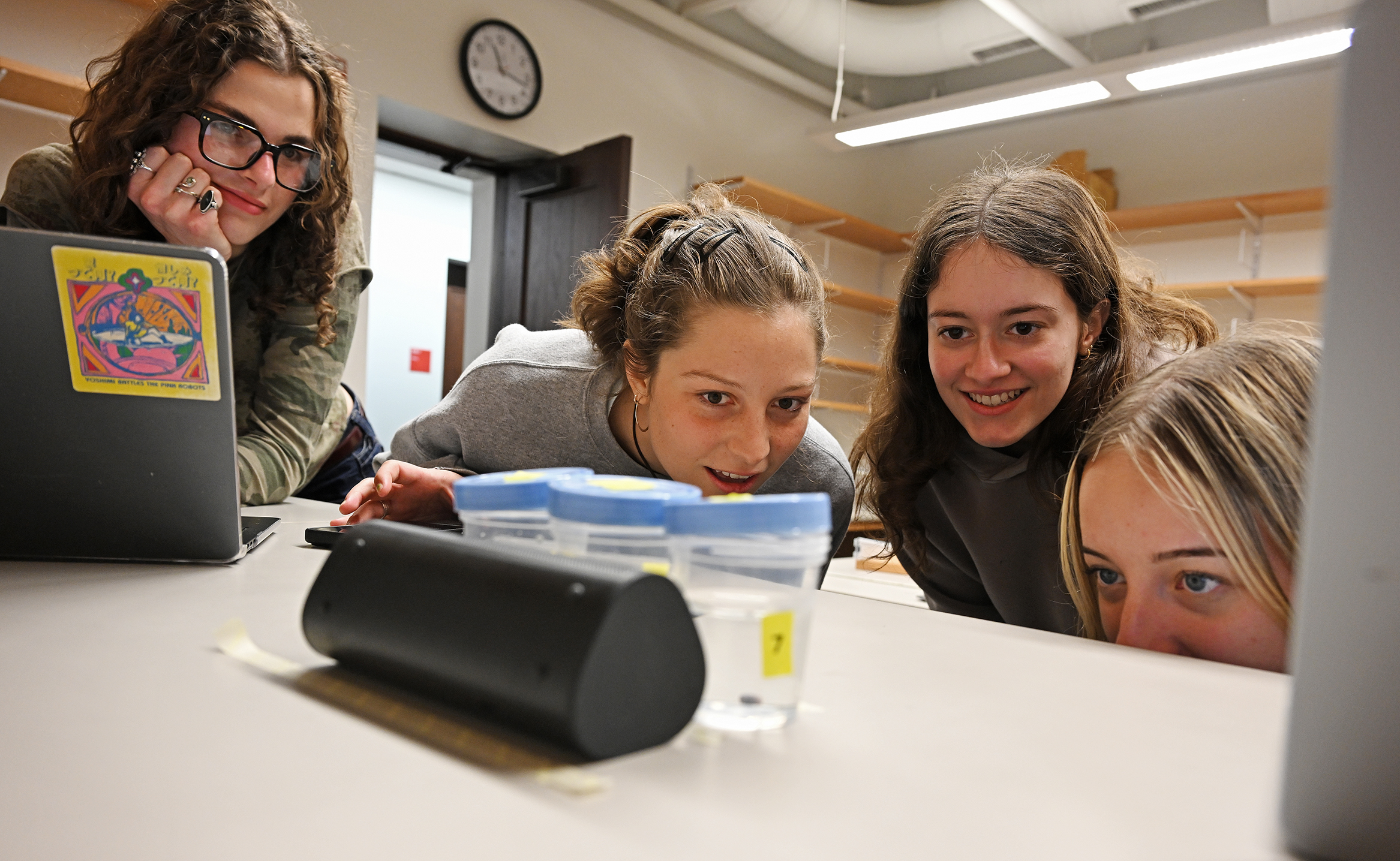 Four female college students gather along a countertop leaning in to look closely at some small containers.