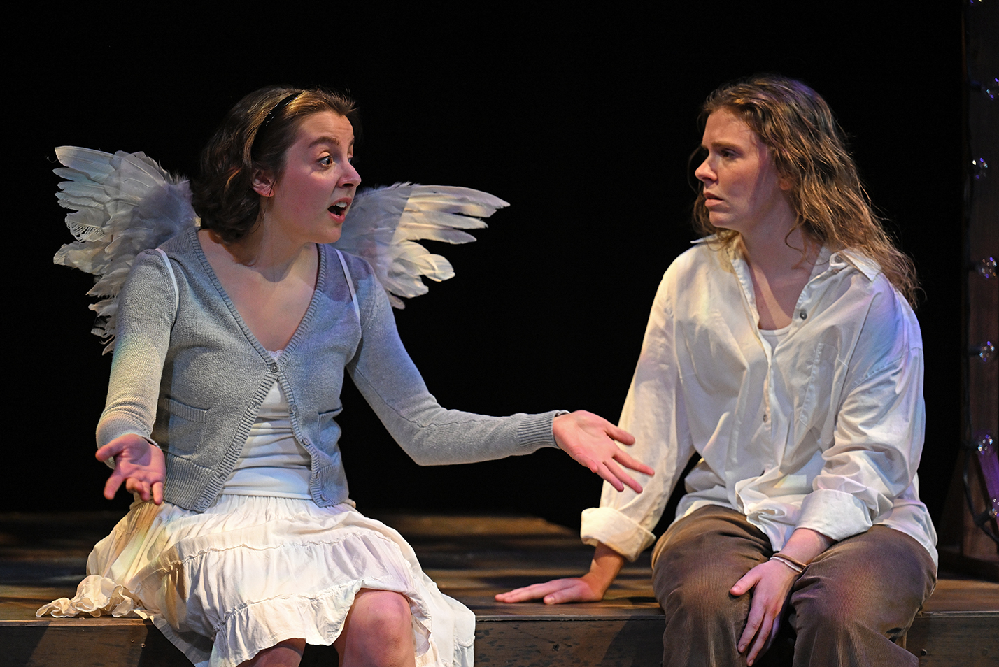 Two seated student actors, one wearing feathered wings and a skirt the other in a loose blouse and khaki pants, act out a scene on stage in a black box theater.