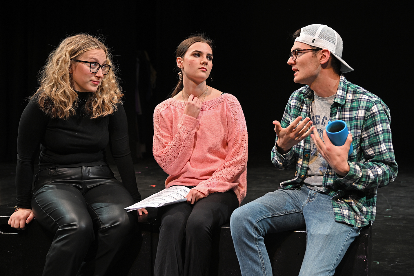 Three student actors, one dressed in all black, one in a pink shirt, and one in a plaid shirt with jeans and a reversed baseball cap, act out a play scene in a black box theater.