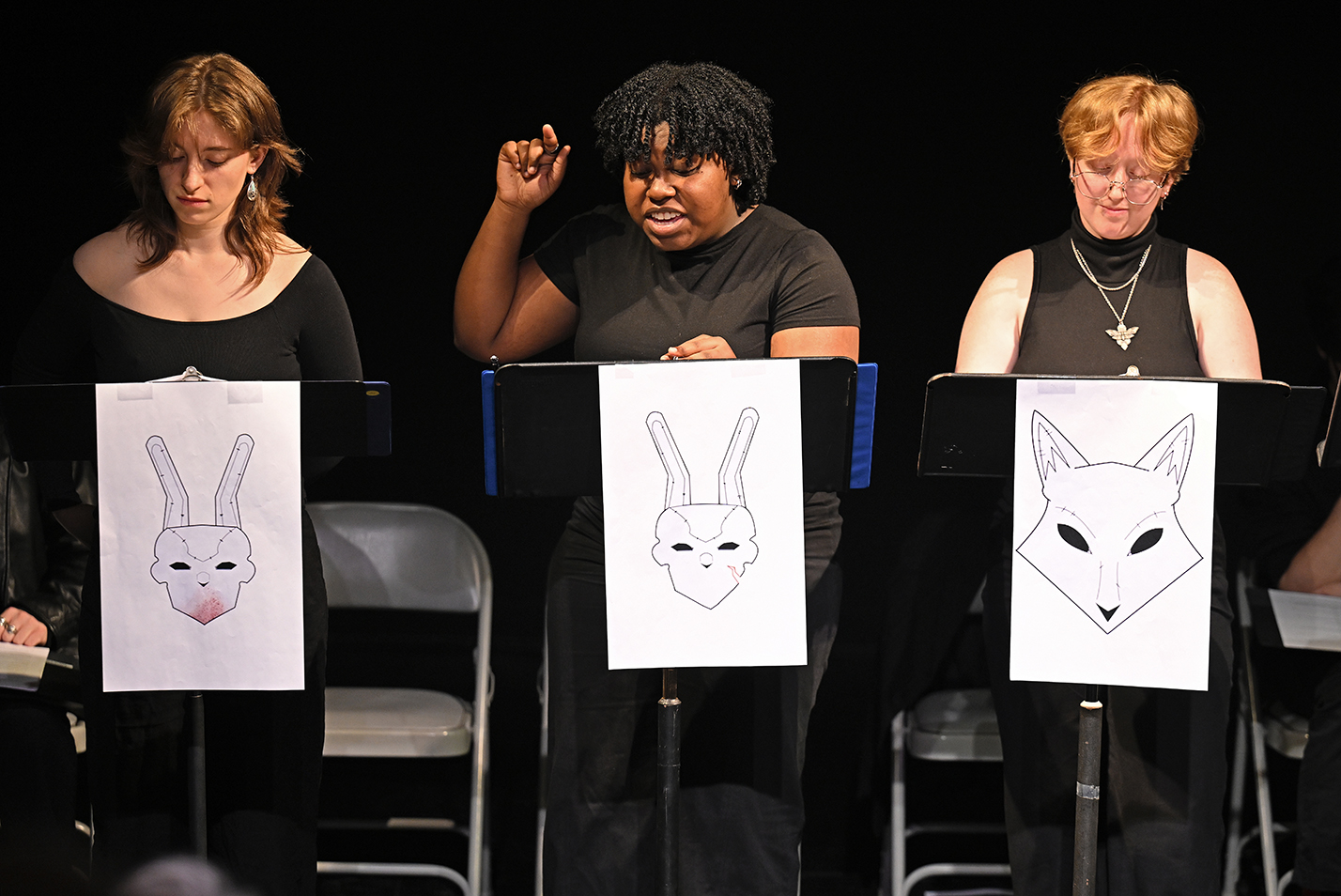 Three student actors all in black t-shirts and standing behind music stands adorned with animal character mask drawings perform a staged reading of a play.