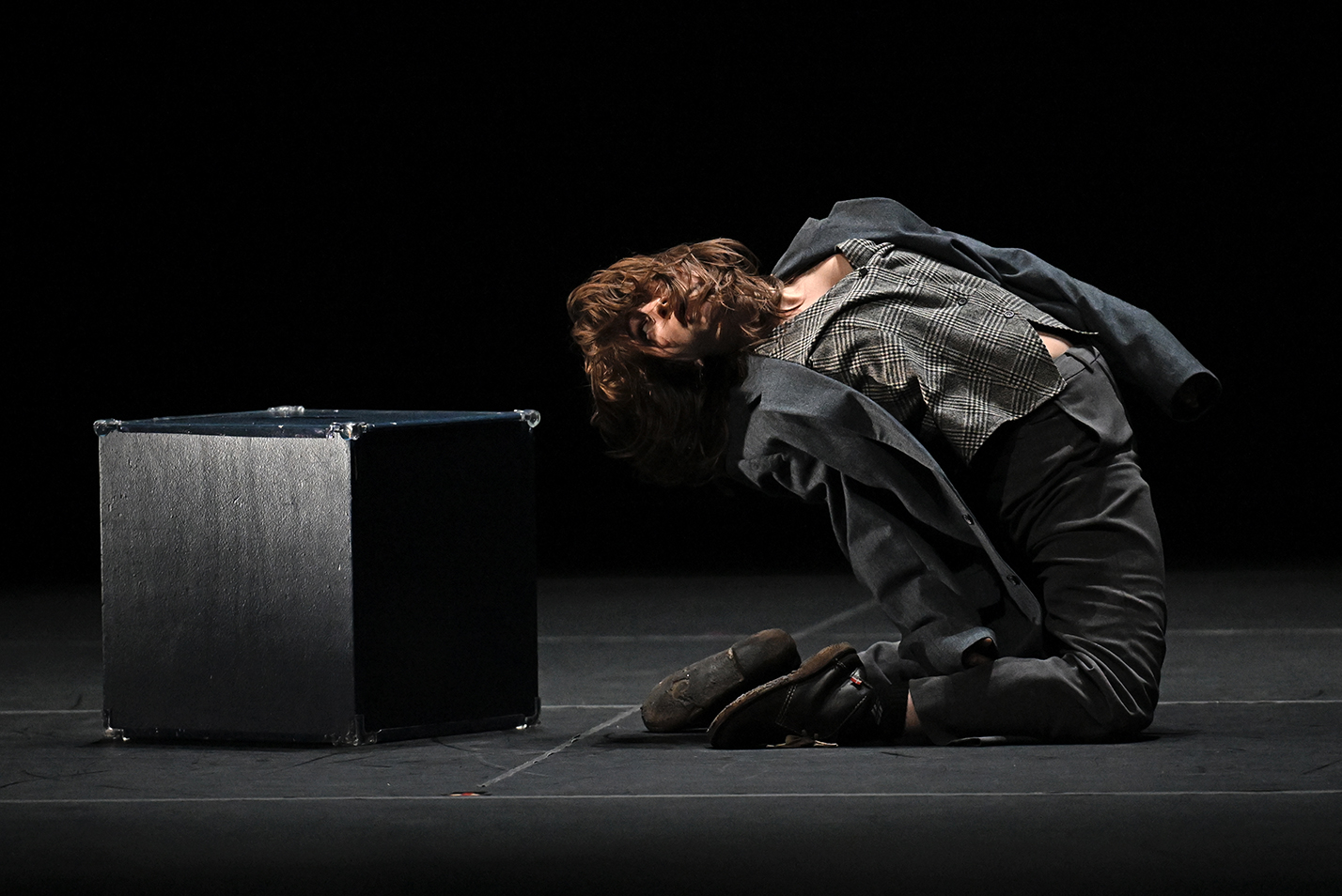A student dancer in rumpled suit bends over backwards while kneeling next to a black box on a stage.