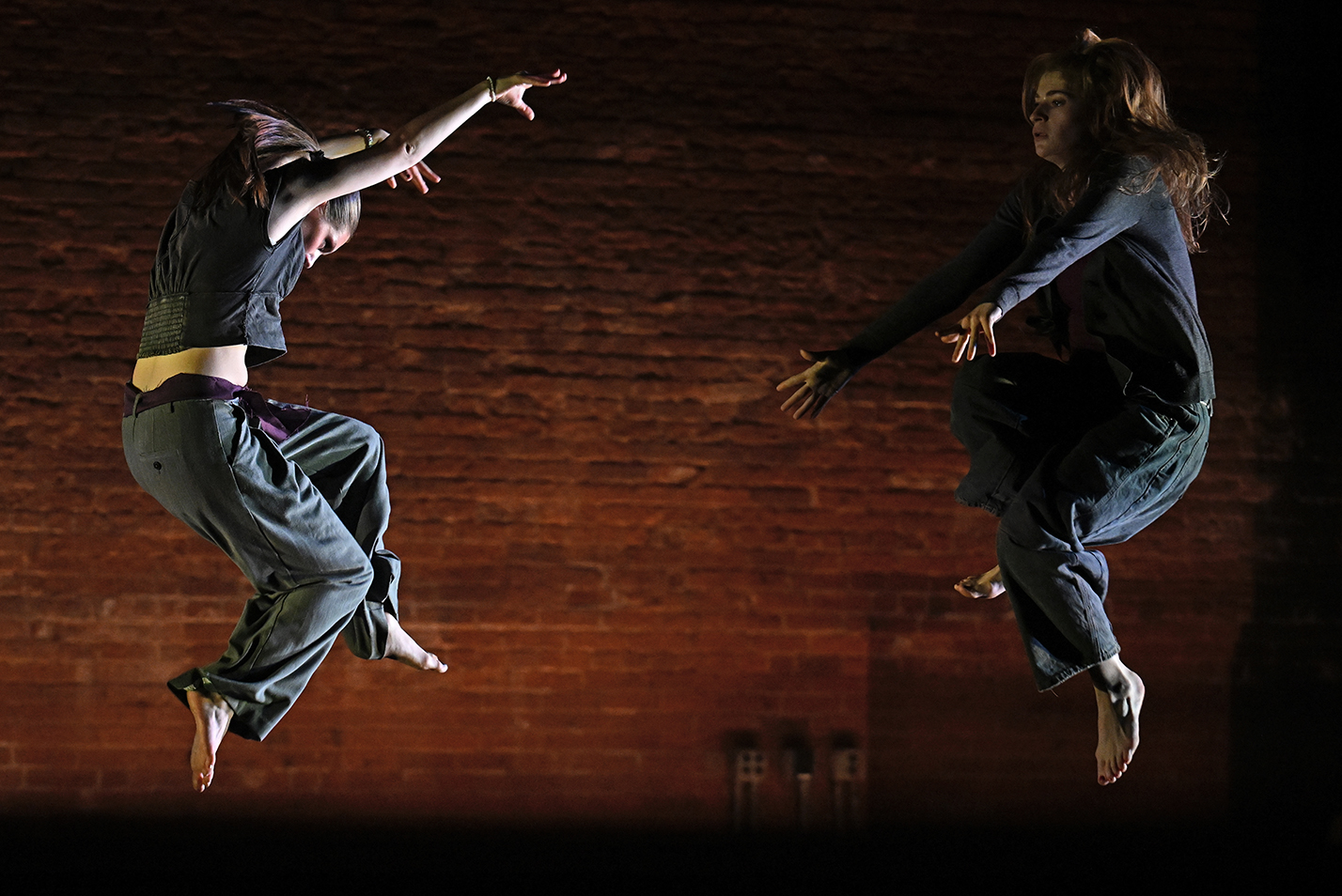 Two dance students leap high off the floor with one reaching up with her arms and the other down with her arms against a backdrop of a brick wall.