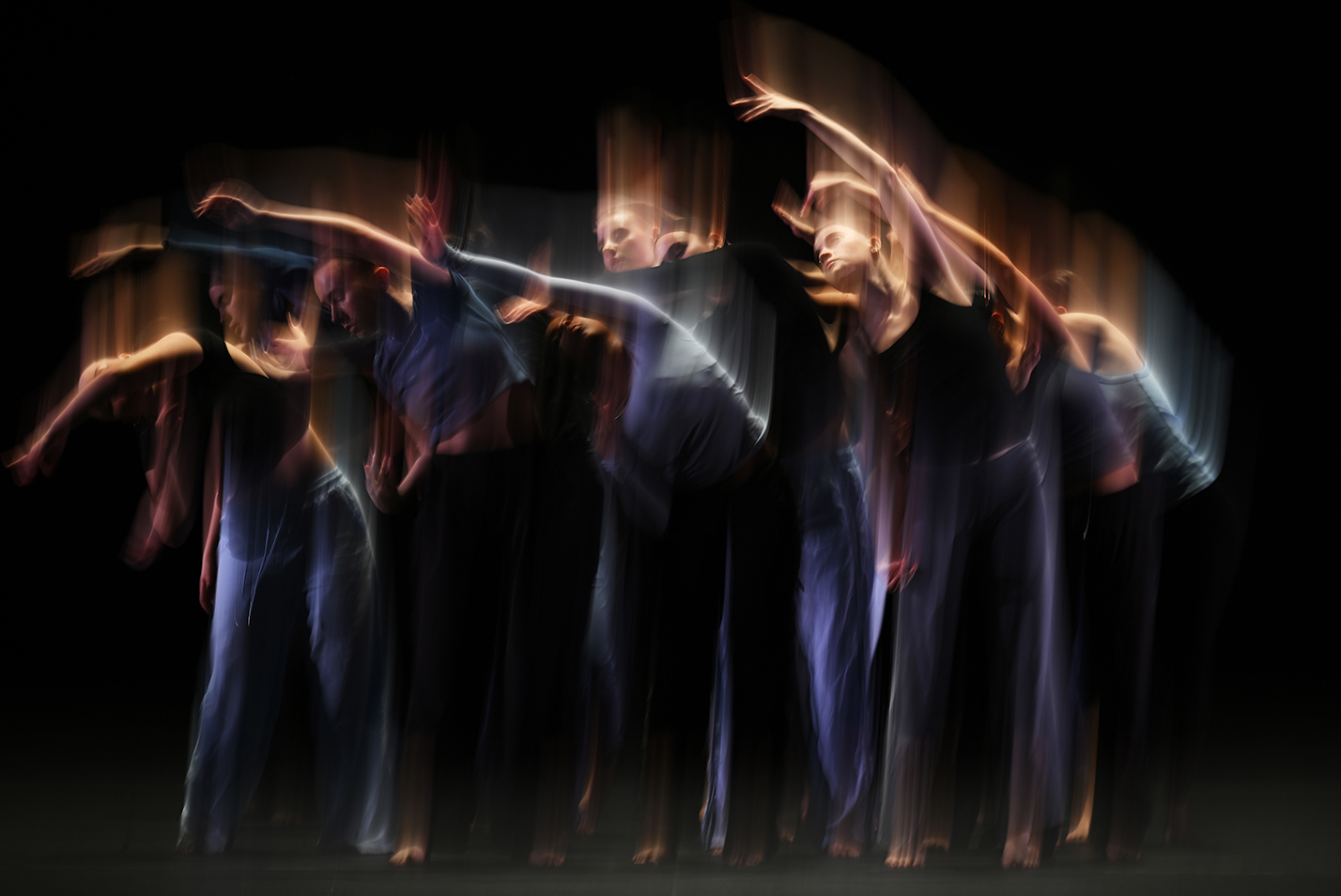 A group of dancers in muted blue tones and shown with vertical motion blur perform a modern dance piece on stage.