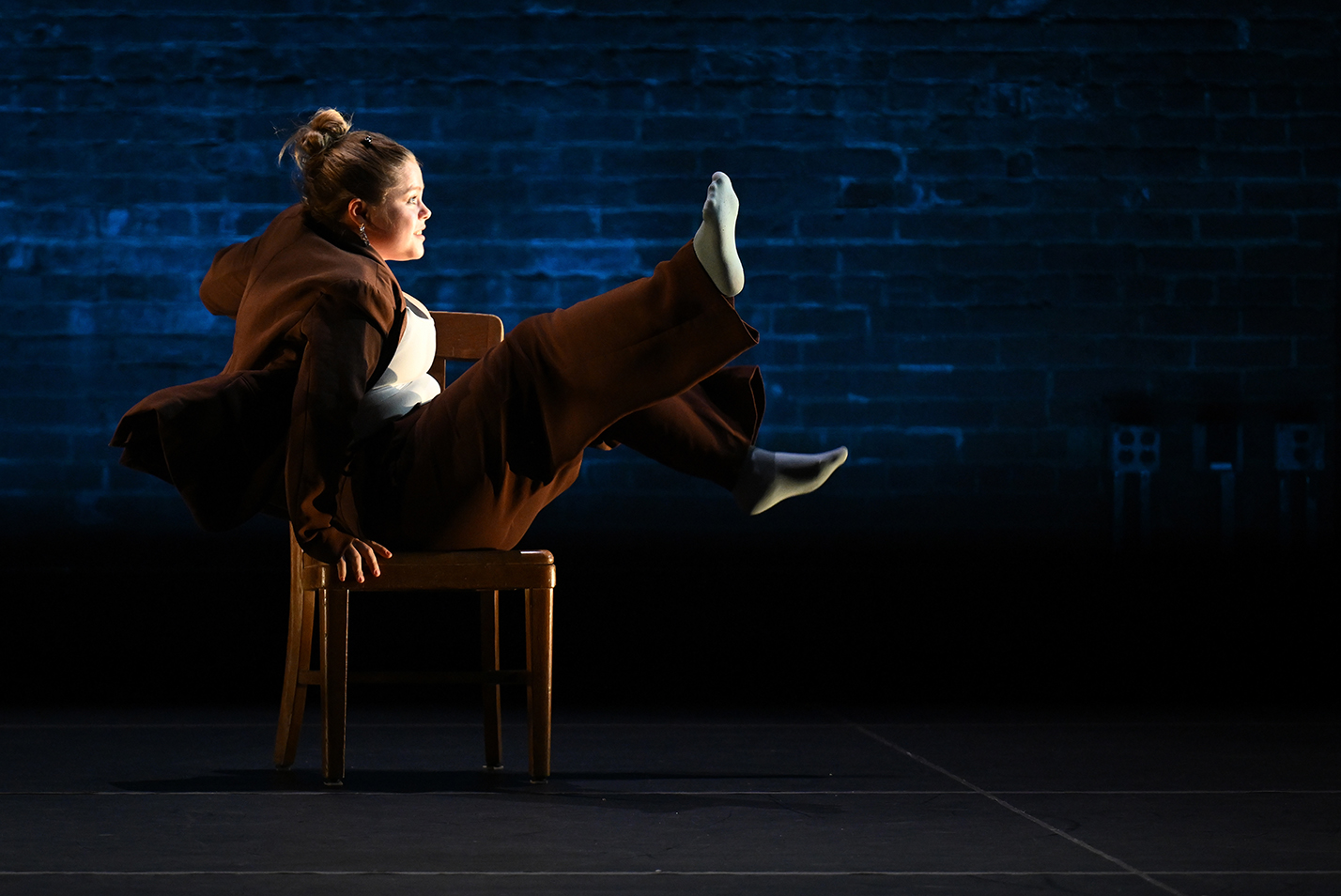 A student dancer in brown pantsuit and hair in a bun throws her legs in the air while seated sideways on a wooden chair in a modern dance piece on a stage with a brick wall in the background.