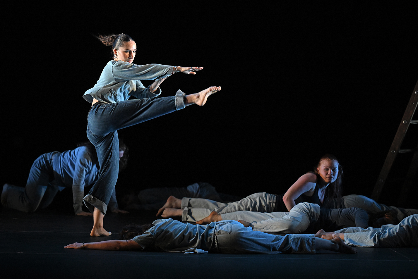 A dancer in blue jeans and a light blue long-sleeve shirt kicks a pointed to our over dancers sprawled across the floor in a modern dance piece performed on stage.