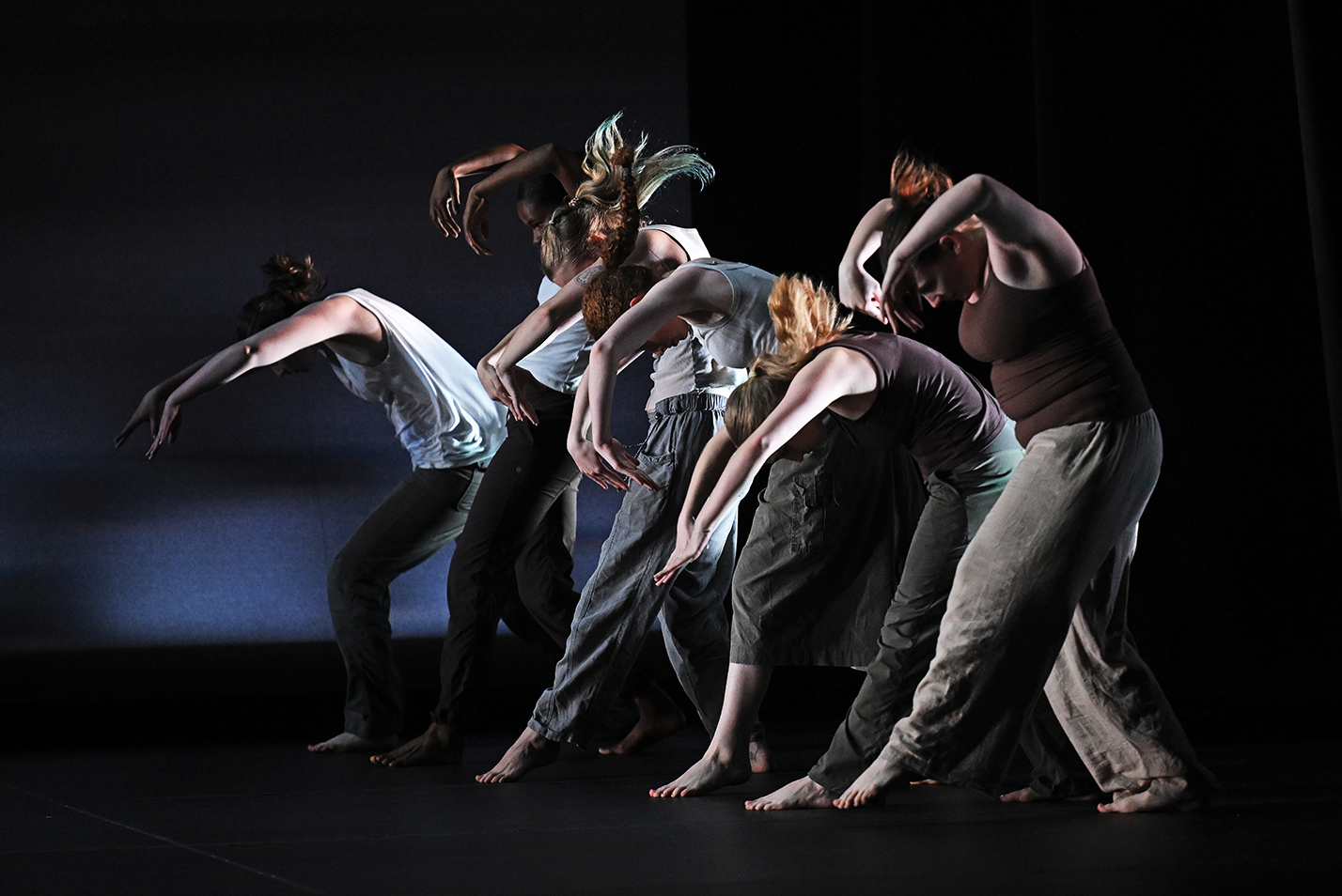 Modern dancers in loose long pants and sleeveless tops create a wave motion with curved arms and arced backs while performing on stage.