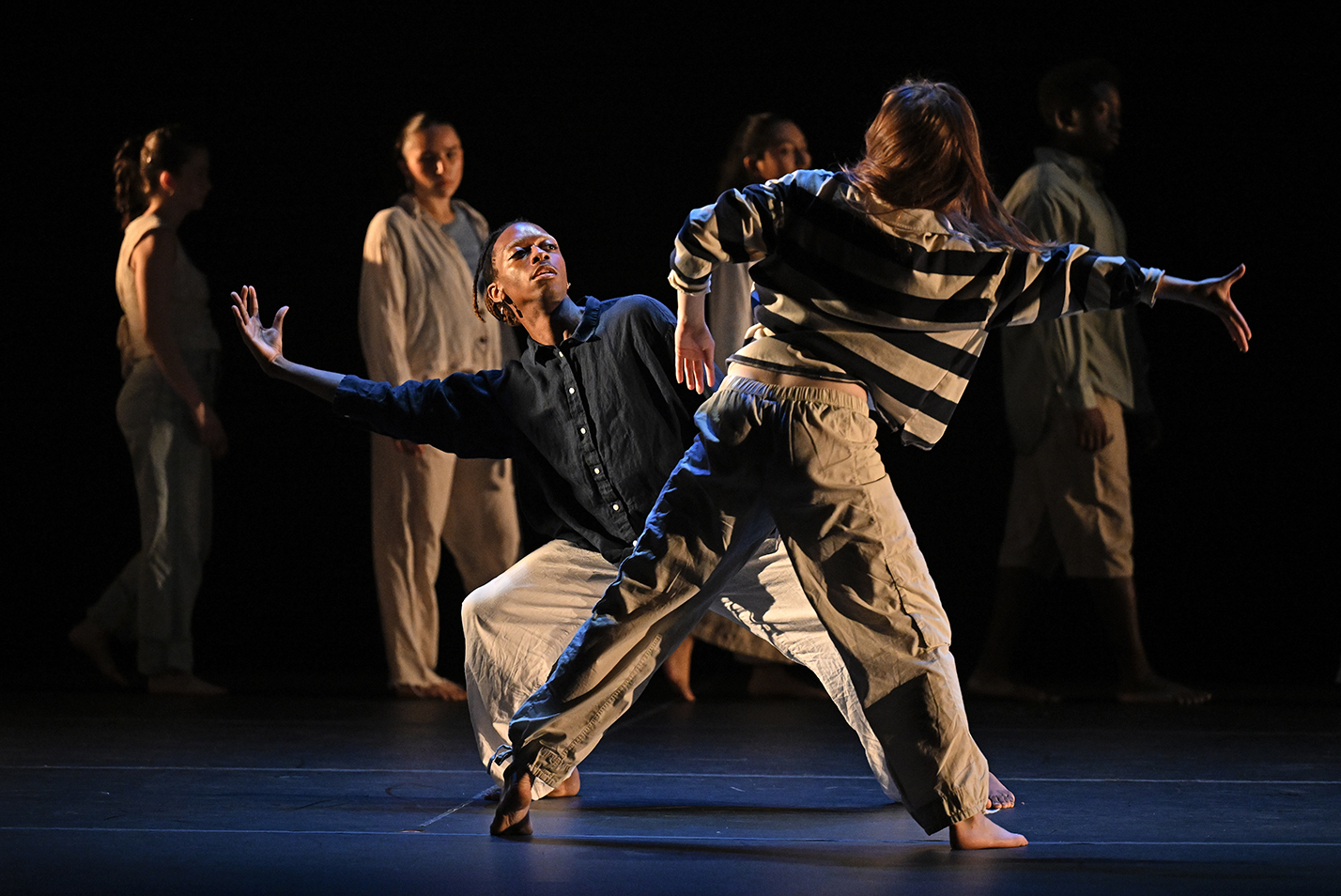 Two dancers, one in a horizontally striped shirt, the other in a dark blue button-down shirt, mirror each others moves while other dancers watch standing in the background.