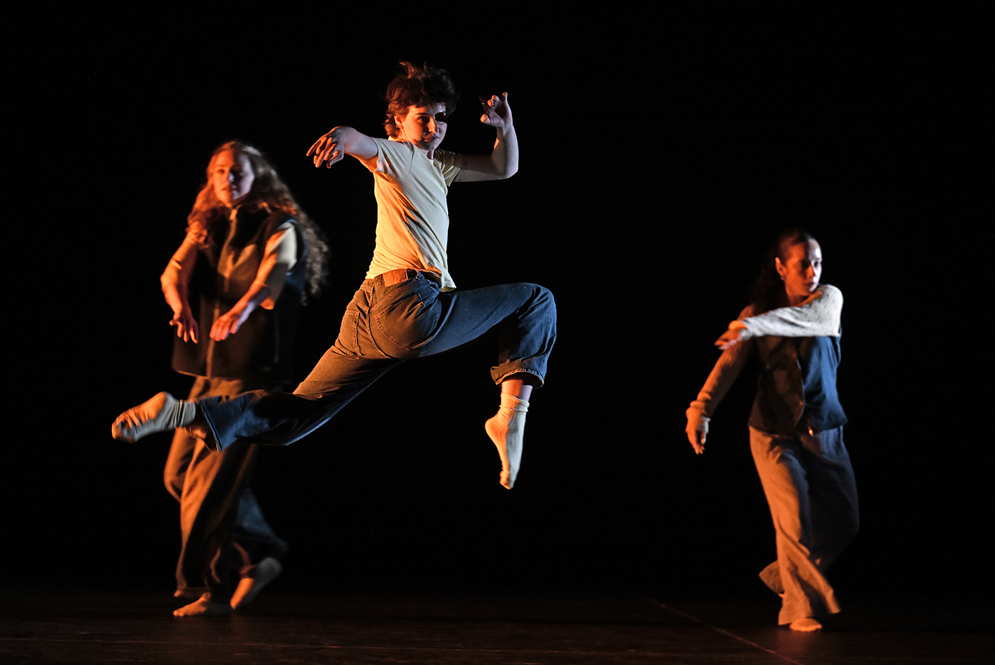 A student dancer leaps high in the air in a hurdling motion while looking back over their shoulder towards the audience while two dancers are in the background.